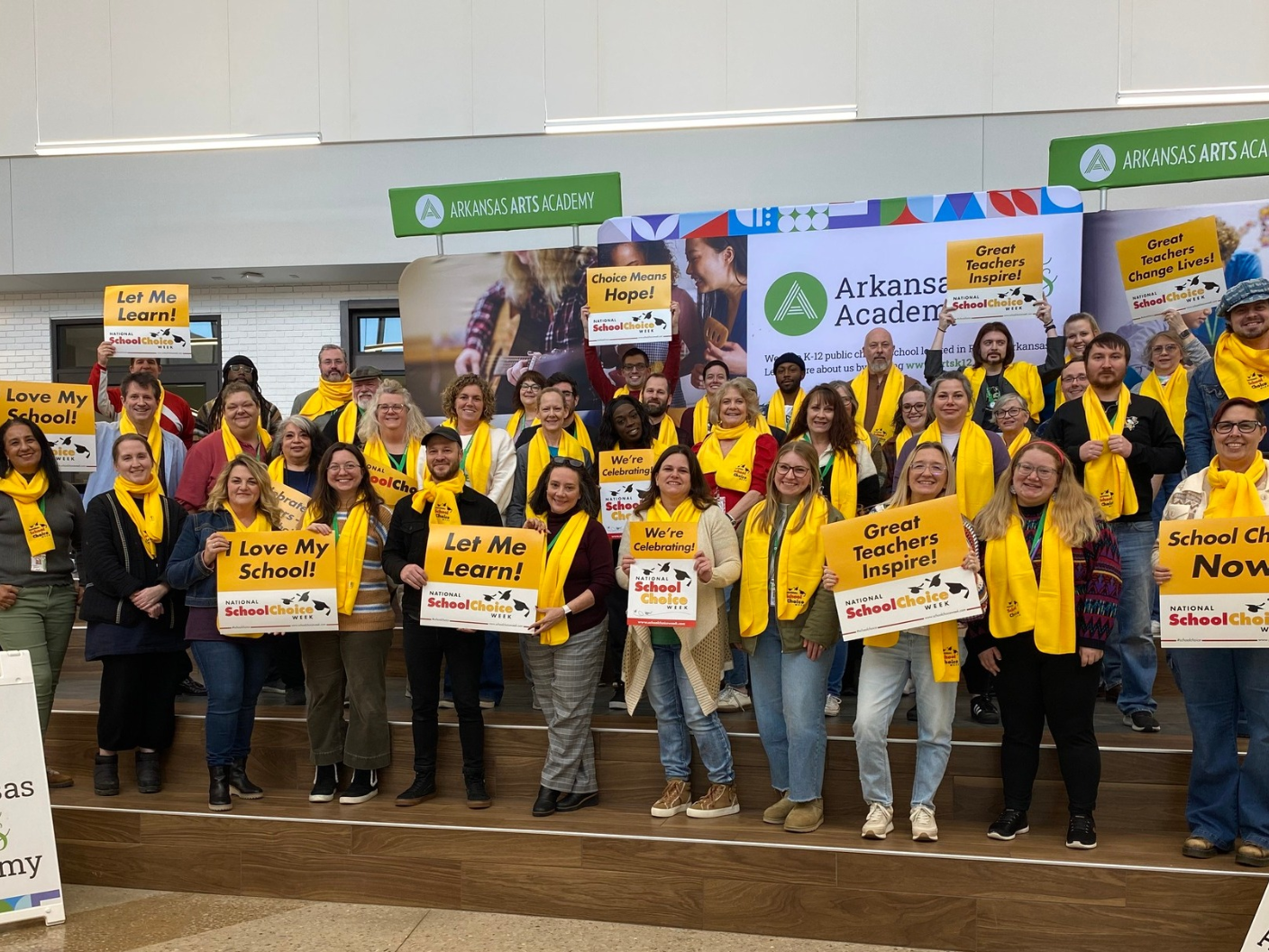 Arkansas Arts Academy staff standing on stage, holding signs and wearing scarves, celebrating National School Choice Week.